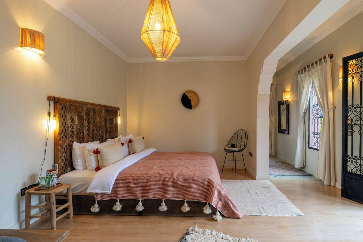A bedroom with a wooden headboard, a bed with a striped blanket, a hanging light fixture, and a view of a hallway with a door.