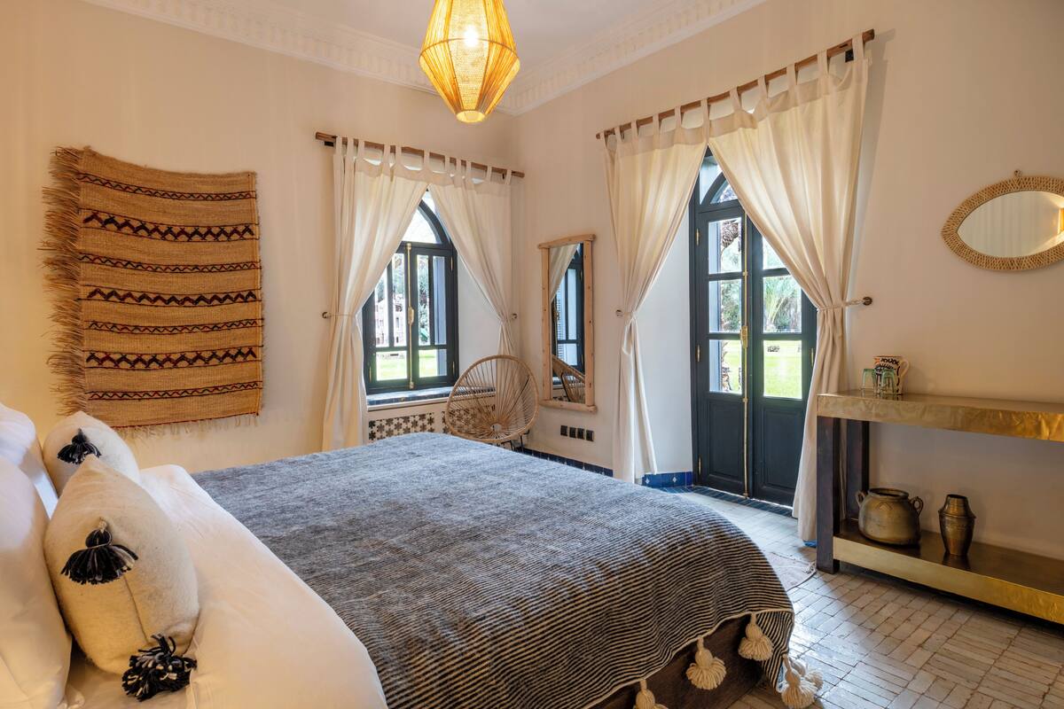 A bedroom with a bed, a chair, a mirror, a shelf with items, and a window with curtains.