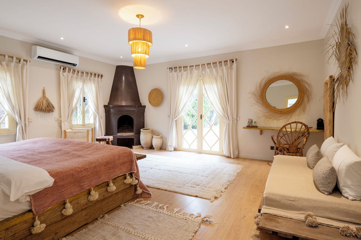 A bedroom with a fireplace, a bed with a red and white striped bedspread, a sofa with white and gray pillows, and a round mirror on the wall.