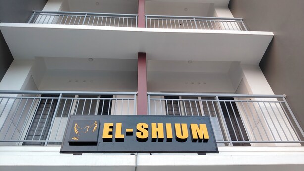 Un immeuble à plusieurs étages, sur lequel on peut lire l’inscription « EL-SHIUM » et qui arbore un logo représentant un oiseau.