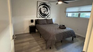 Ein Schlafzimmer mit Bett, Deckenventilator, Wandbild, Nachttisch und Fenster mit Jalousien.