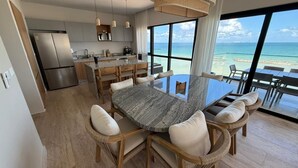 Et moderne køkken med et rustfrit stål køleskab, træskabe og en stor ø med siddepladser. Et spisebord med stole står i midten, og et stort vindue giver udsigt til havet.
