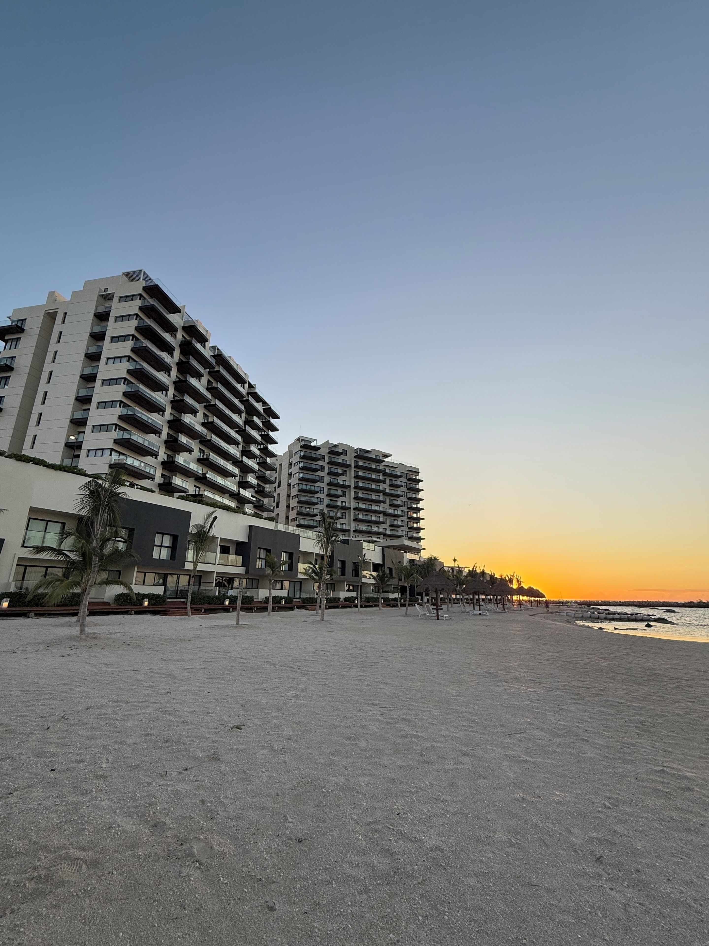 Un hôtel situé en bord de mer, avec plusieurs étages, des espaces de détente en plein air et une vue sur l’océan au coucher du soleil.