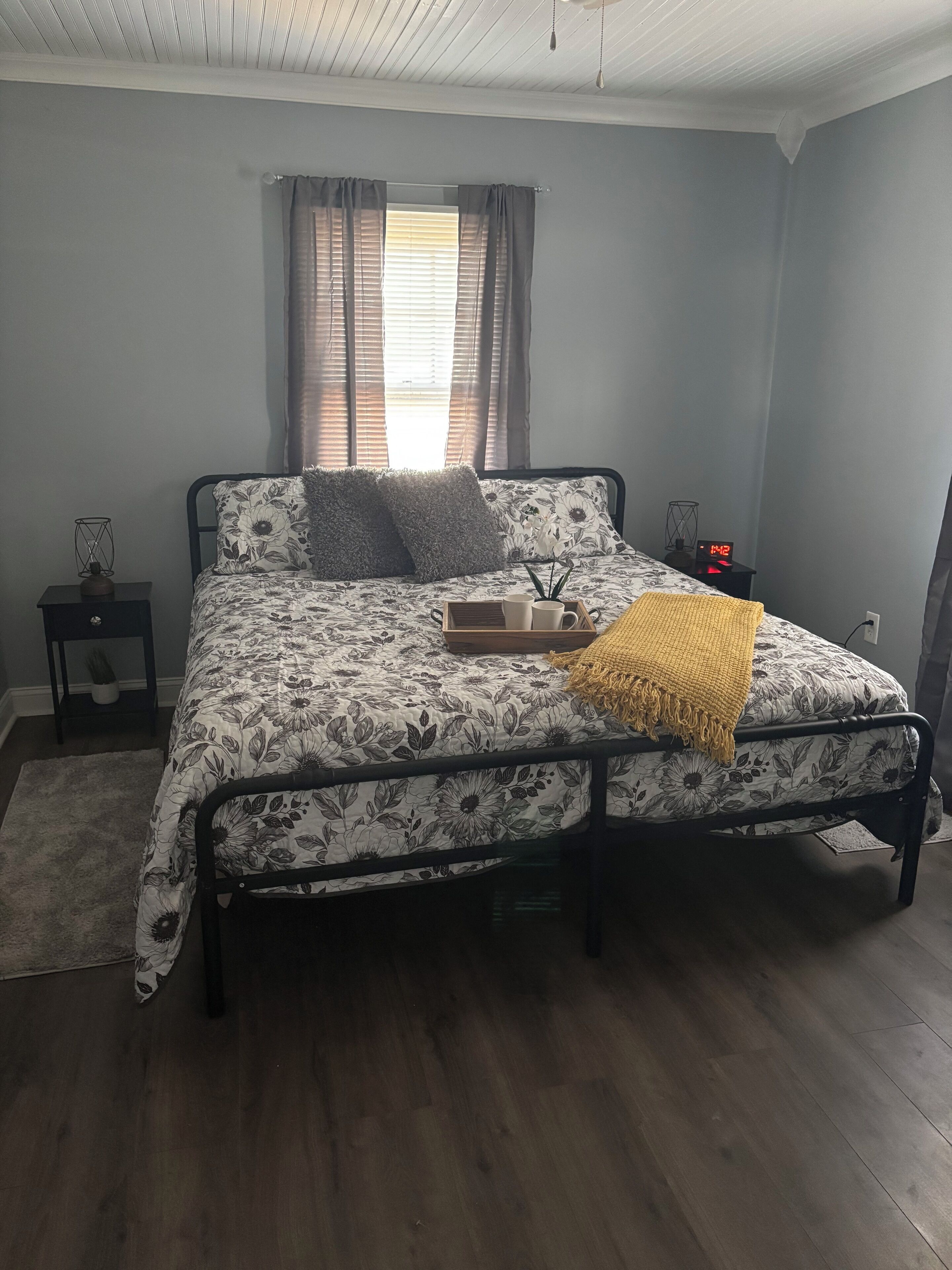 A bedroom with a bed, a bedside table, a nightstand, a window with curtains, and a wall clock.
