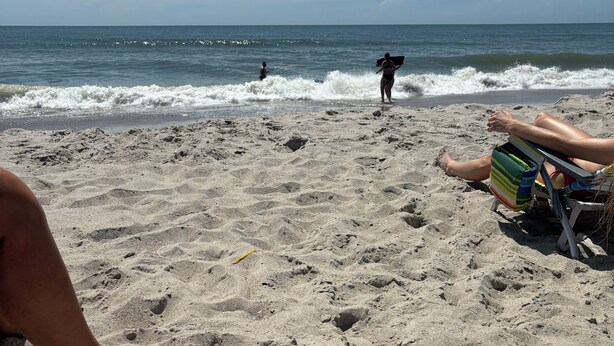 Une plage de sable avec des empreintes de pas, une personne allongée et une autre assise, tenant une planche de surf.
