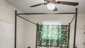 Una cama con dosel, un ventilador de techo, dos sillones tapizados, un sendero estampado y una pared con follaje verde.