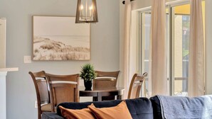 Ein Wohnzimmer mit einem dunklen Sofa, einem Holzespeisetisch mit Stühlen, einer Hängeleuchte und einem großen Fenster mit Vorhängen.