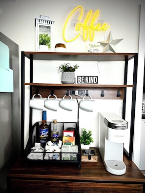 Una estación de café con una máquina Keurig, un cartel que dice 'Coffee is my true love' y un letrero que reza 'Be Kind' sobre una estantería.