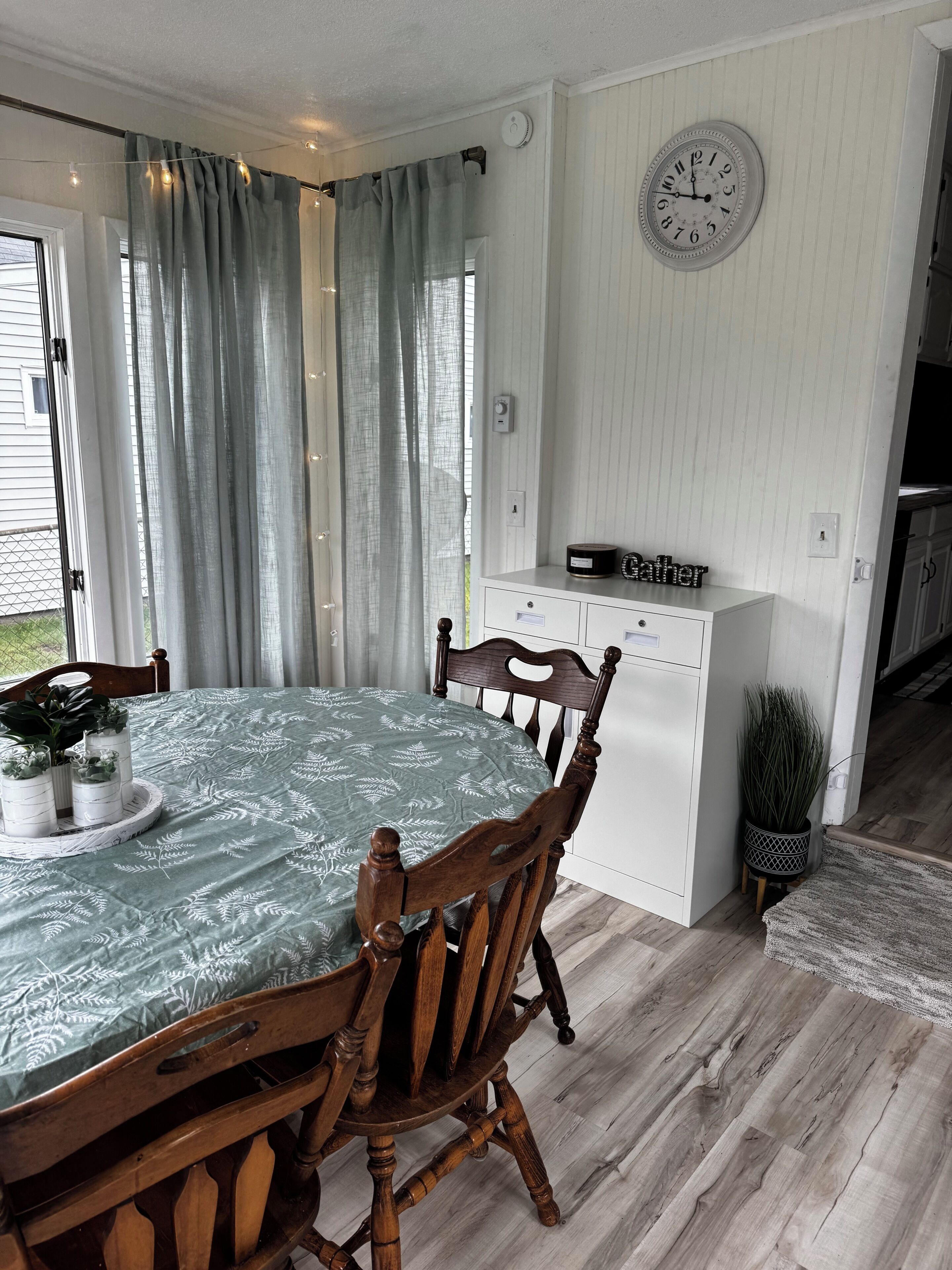 A dining area with a table set for two, wooden chairs, a clock on the wall, and a window with curtains.