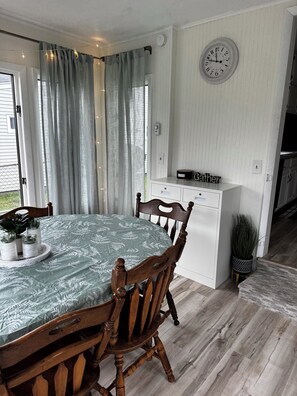 Uma área de refeições com uma mesa posta para dois, cadeiras de madeira, um relógio na parede e uma janela com cortinas.