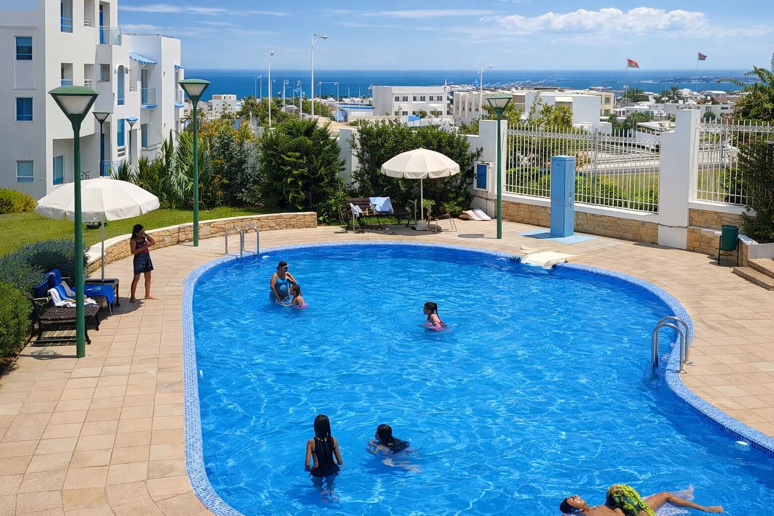 A swimming pool with people, a view of the sea, and buildings in the background.