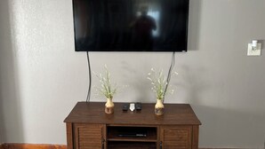 Un téléviseur à écran plat fixé au mur, au-dessus d’un meuble en bois servant de support pour l’équipement audio-vidéo, et agrémenté de deux petits vases de fleurs.