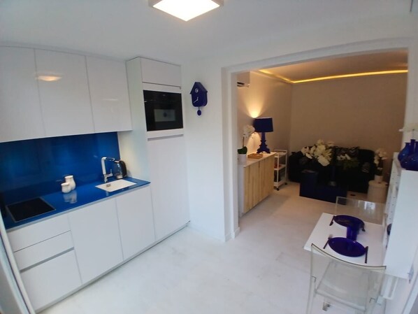 A modern kitchen with white cabinets, a blue countertop, and a built-in oven. There is a small dining area with a white table and blue chairs.