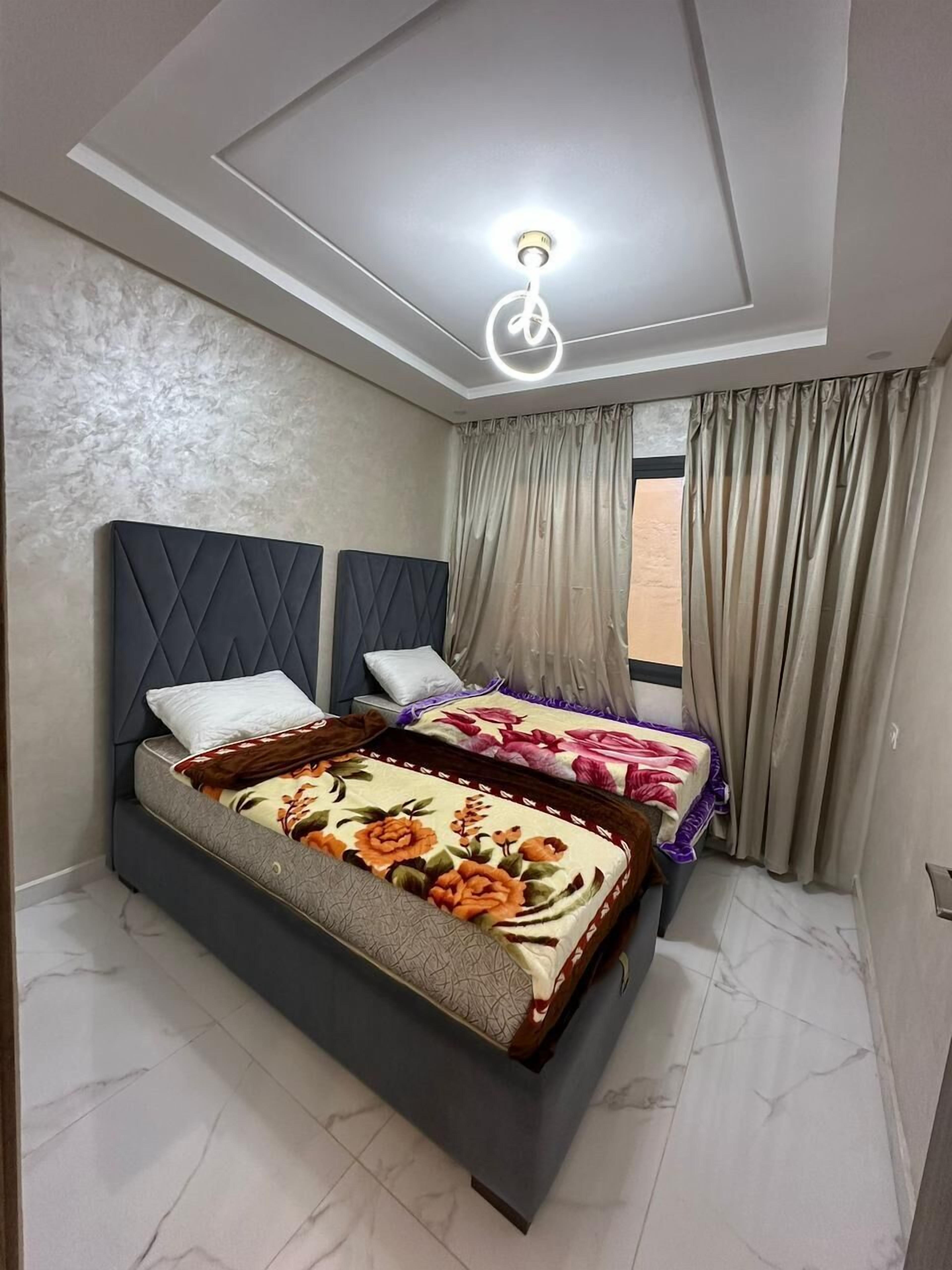 Une chambre à coucher avec un lit, deux têtes de lit, un luminaire au plafond et une fenêtre avec des rideaux.