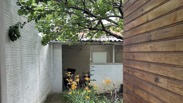 Un étroit couloir extérieur, avec un mur en bois d’un côté et un mur en briques blanches de l’autre. On y trouve des plantes en pot et un petit espace jardin agrémenté de fleurs jaunes.