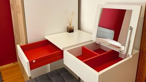 Ein kleines Zimmer mit weißem Schreibtisch, einer roten Schublade, einem Spiegel und einem kleinen Regal mit einem Schilferdiffusor.