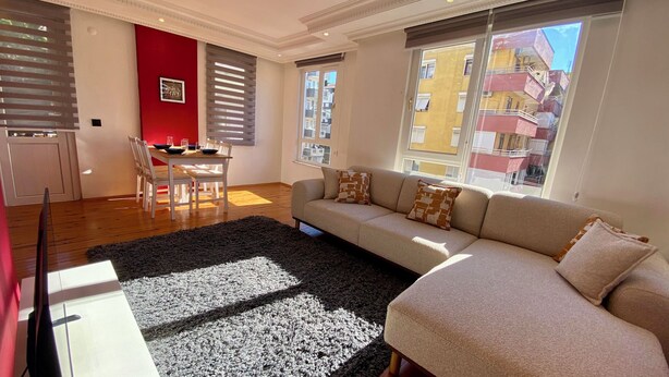 Ein Wohnzimmer mit einer Couch, einem Esstisch mit Stühlen, einer roten Akzentwand und großen Fenstern mit Blick auf Gebäude.
