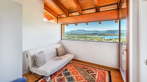 A room with a wooden ceiling, a white sofa, a patterned rug, and a large window offering a view of mountains and a lake.