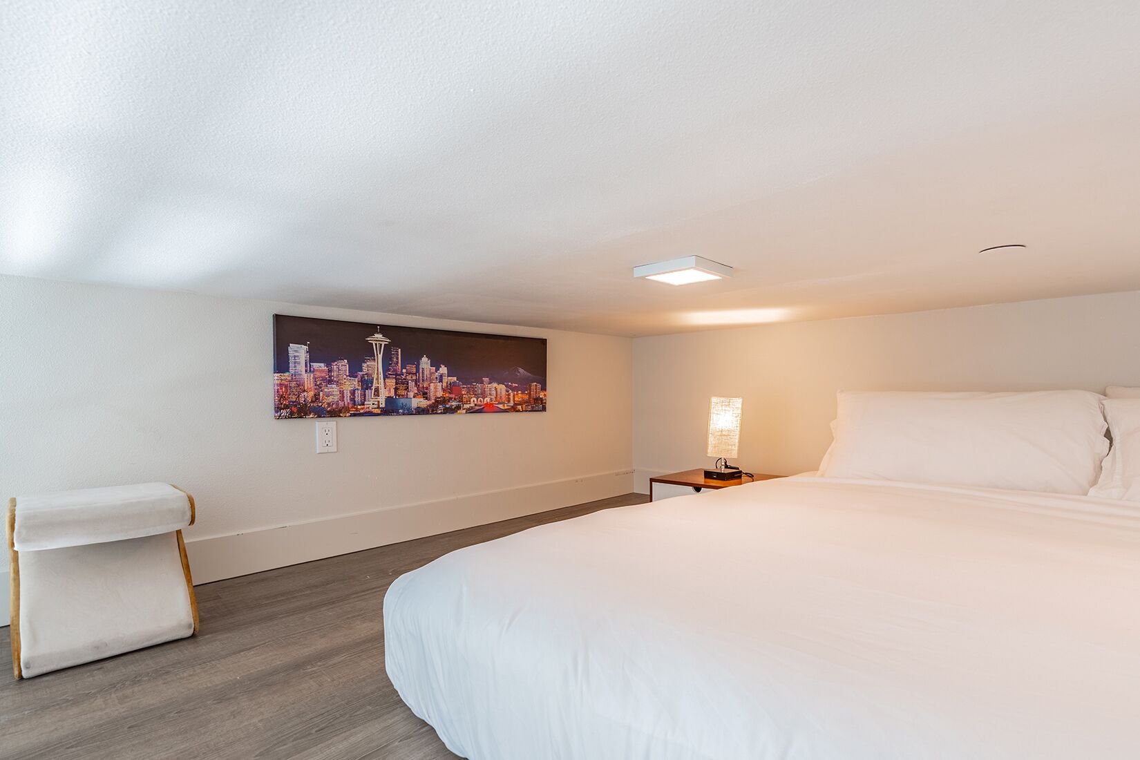 A hotel room with a large bed, a nightstand, a lamp, a chair, and a cityscape painting on the wall.