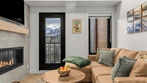 Ein modernes Wohnzimmer mit Kamin, einer Couch, einem Couchtisch mit Obst und Blick auf Berge durch eine Tür.