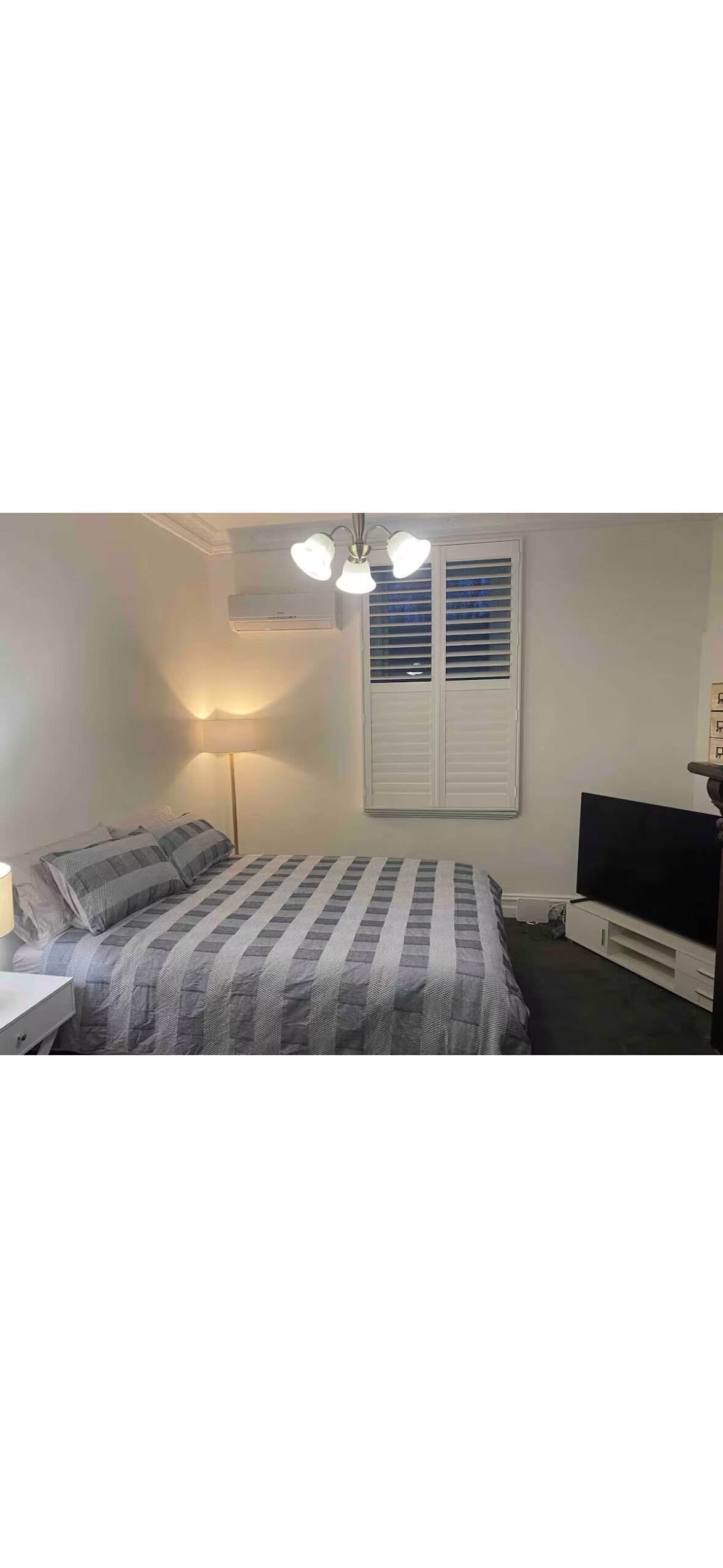 Un dormitorio con cama, lámpara de noche, aire acondicionado, ventana con persianas y un mueble para televisor.