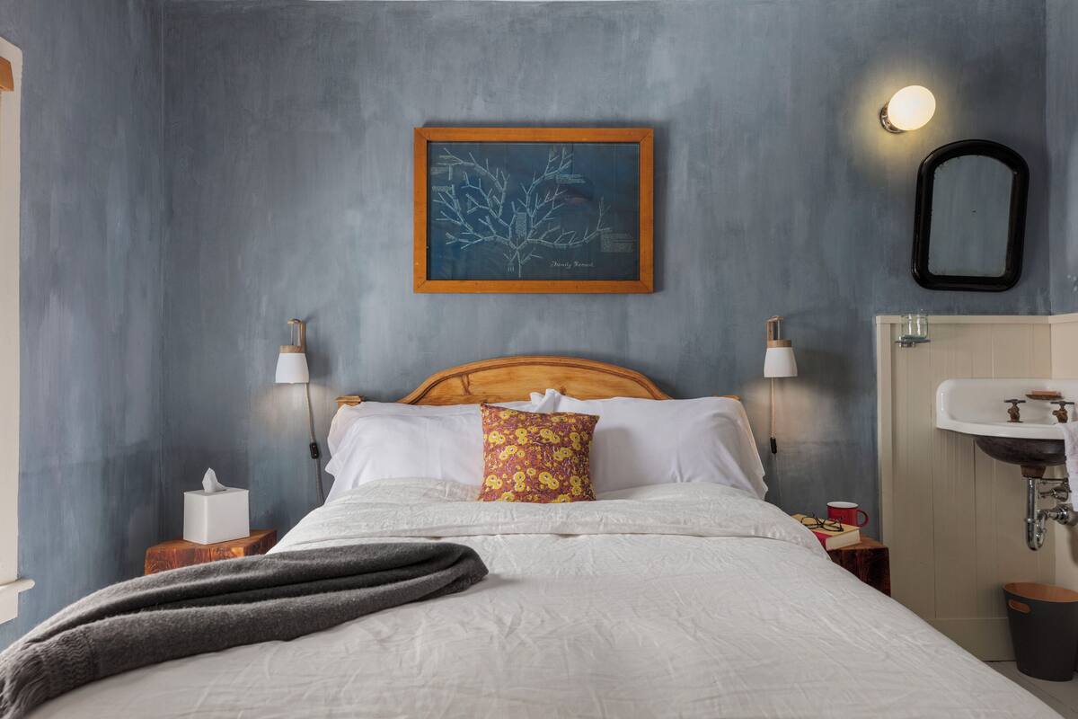 Un letto rifatto con lenzuola bianche, un cuscino colorato e una coperta grigia. Sopra il letto, un quadro incorniciato al muro, una lampada da comodino e un piccolo comodino in legno con una scatola di fazzoletti e una tazza rossa.