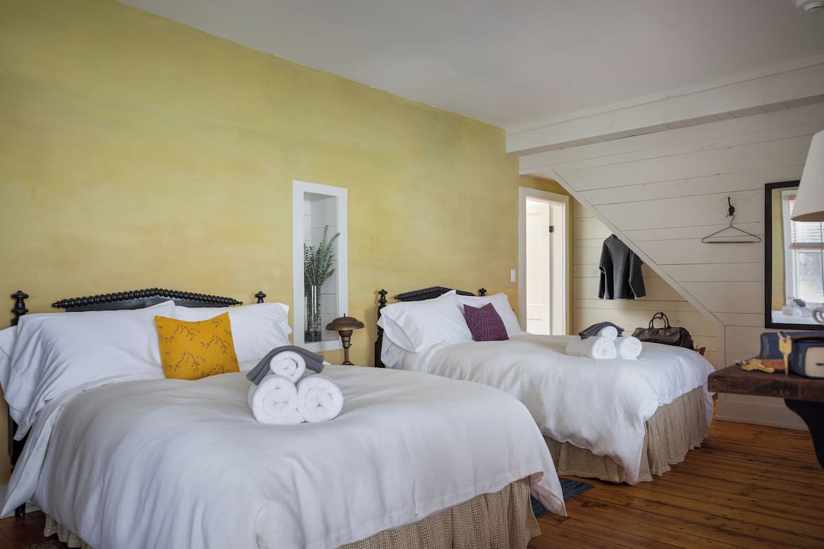 Camera d'albergo con due letti, ognuno con biancheria da letto bianca e un cuscino giallo. Il pavimento è in legno e sulla parete c'è uno specchio.