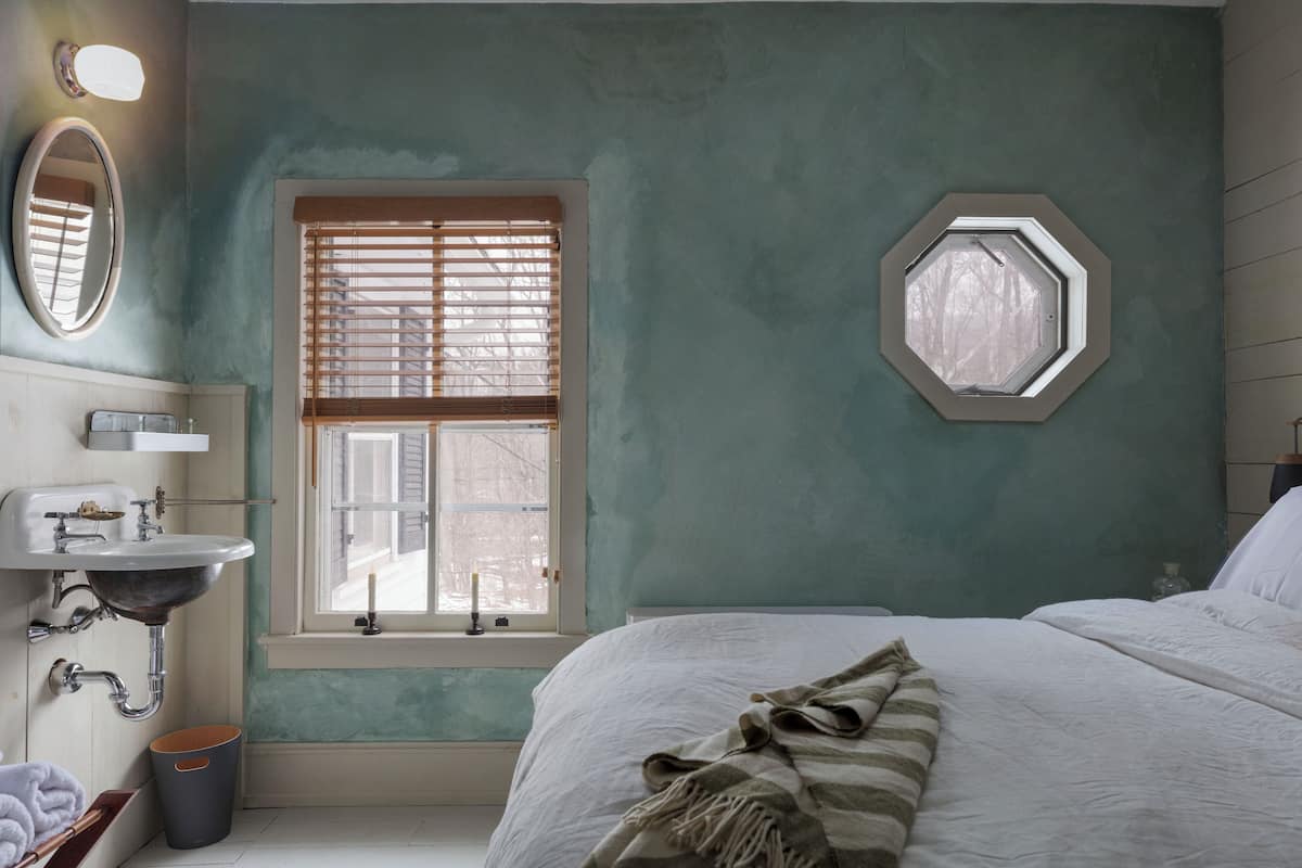 Una camera da letto con un letto, una finestra con tende, un comodino, uno specchio e un lavandino.