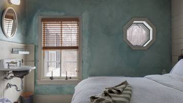 A bedroom with a bed, a window with blinds, a bedside table, a mirror, and a sink.