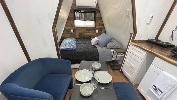 Een ruimtelijk RV-interieur met een blauwe bank, een eettafel met borden en glazen, een kitchenette met een fornuis, en een bed met een raam erboven.