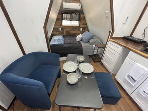 A compact RV interior with a blue sofa, a dining table with plates and glasses, a kitchenette with a stove, and a bed with a window above.