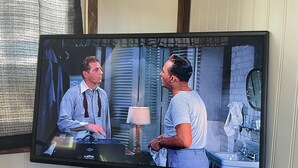 Een televisie toont een scène met twee personages in een kamer met een wastafel en spiegel.