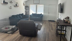 Ein Wohnzimmer mit Ecksofa, einem Sessel, einem Fernseher auf einem Schrank und einem Bücherregal.