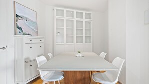Een moderne eetruimte met een witte tafel, vier witte stoelen, een houten kast en een groot raam met uitzicht op het strand.