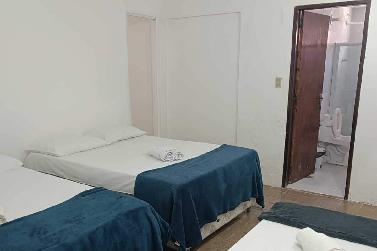 Une chambre d’hôtel avec trois lits simples, une salle de bain visible à travers une porte ouverte et un sol carrelé.