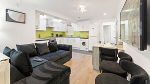 A modern living room with a black sofa, a dining table with chairs, a kitchen with white cabinets, and a large TV mounted on the wall. - London 3 Bedroom Cosy Clean Home Sleeps 6 Stratford Excel Modern (London)