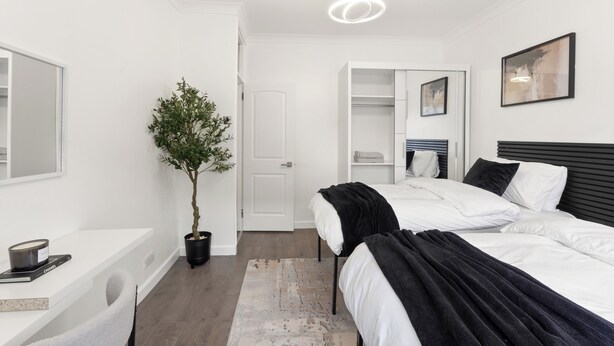 A modern hotel room with two beds, a desk, a potted plant, and a large mirror. - London 3 Bedroom Cosy Clean Home Sleeps 6 Stratford Excel Modern (London)