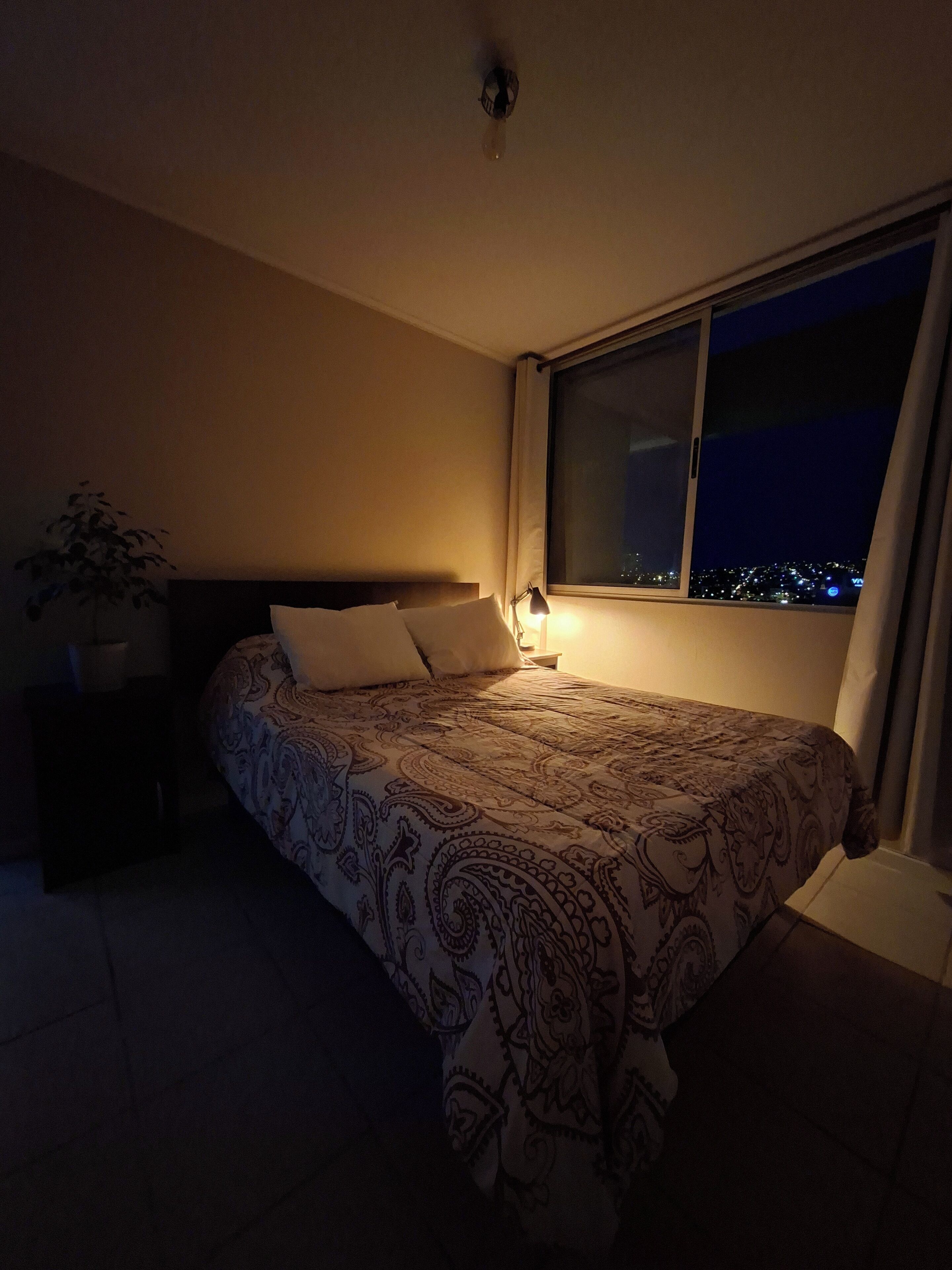 A bedroom with a large bed, a nightstand, a lamp, and a window overlooking a cityscape at night.