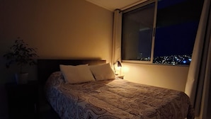Una camera da letto con un letto grande, un comodino, una lampada e una finestra che si affaccia su una vista notturna della città.