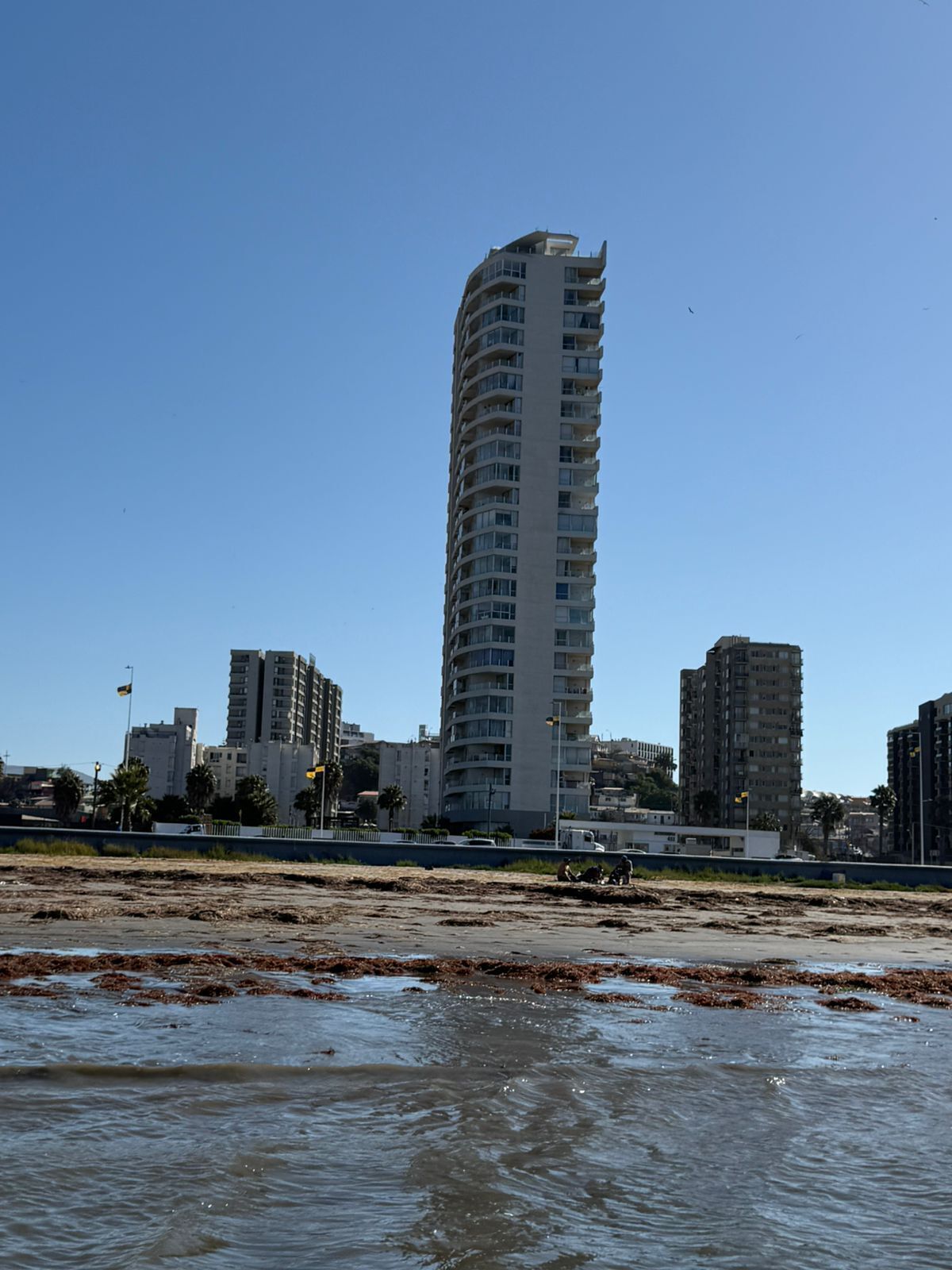 A tall, modern building with multiple balconies, situated near a beach with sandy terrain and water.