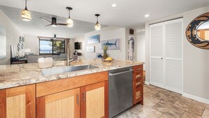 Una cocina moderna con encimeras de granito, armarios de madera, lavavajillas de acero inoxidable, luces colgantes y vista al área de estar.