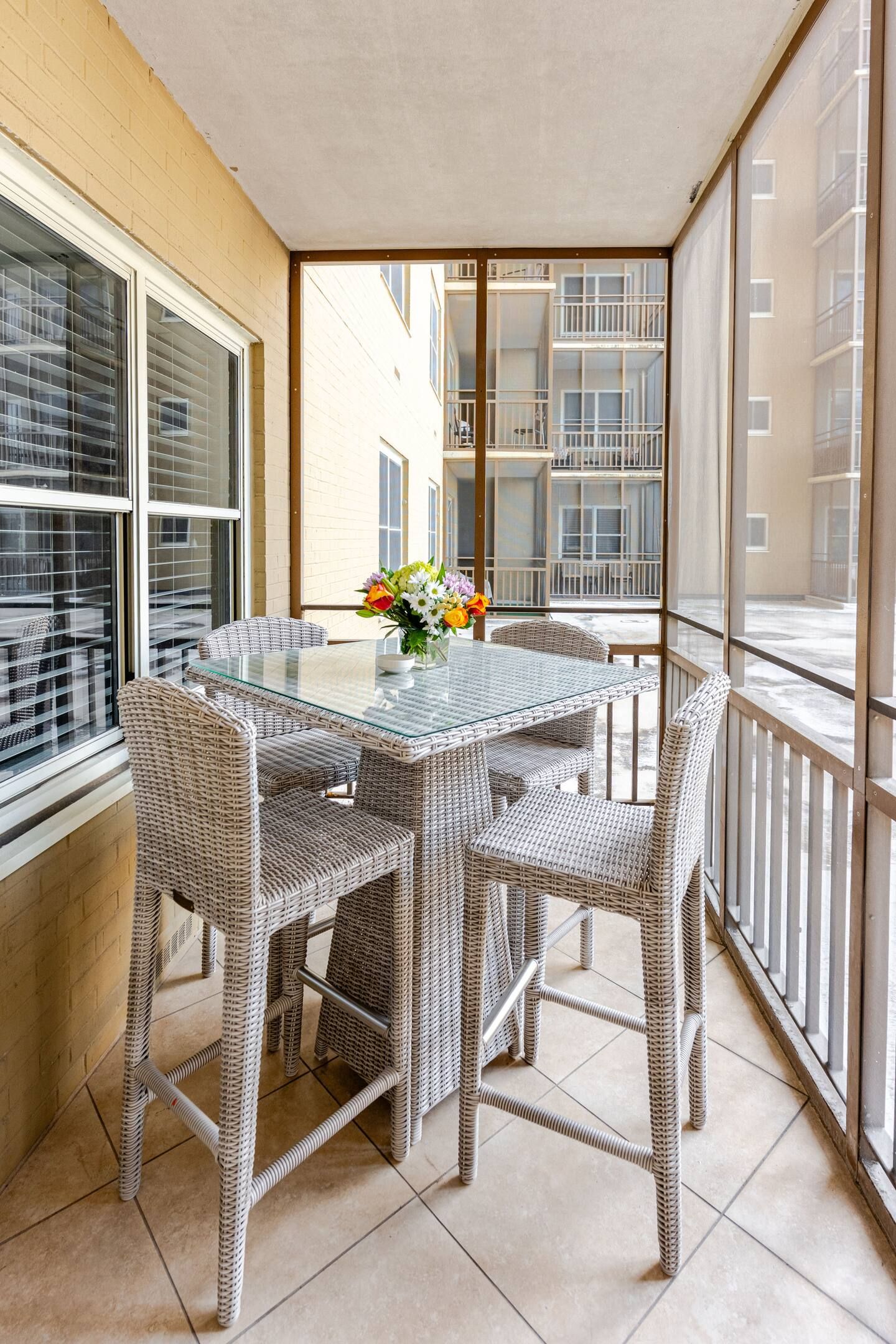 Un balcon doté de meubles en osier et d’une table en verre, offrant une vue sur un immeuble comportant de nombreux balcons.