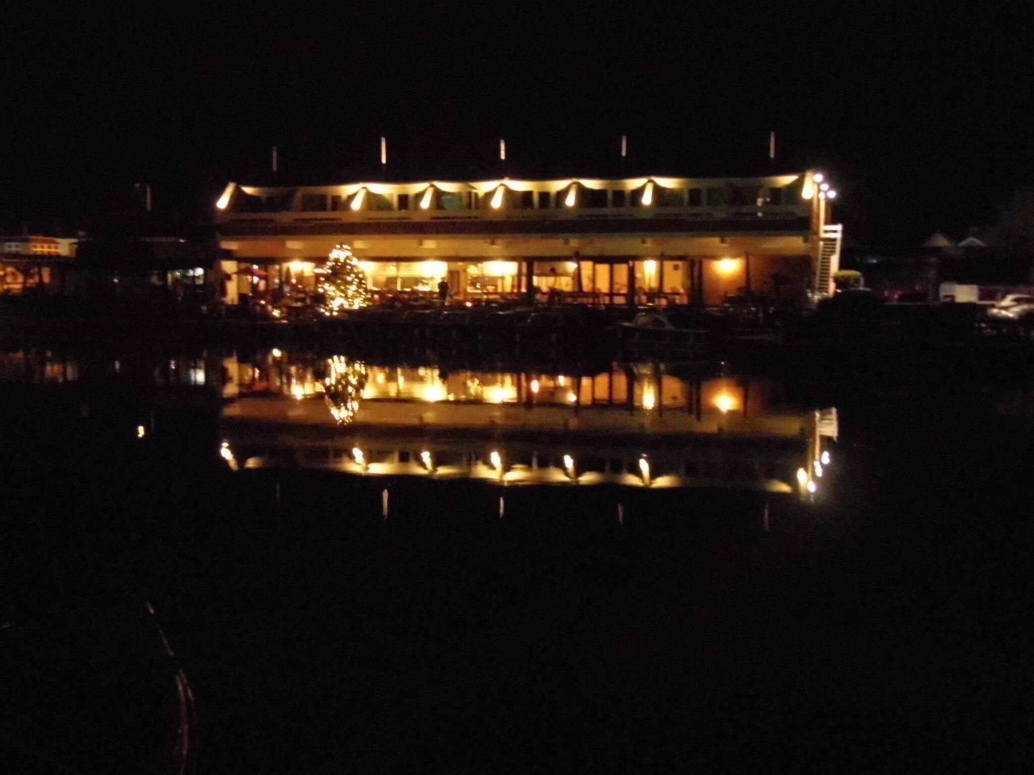 Un bâtiment situé au bord de l’eau, dont l’image se reflète dans l’eau, avec un grand sapin de Noël illuminé et de nombreuses fenêtres.
