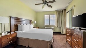 A hotel room with a large bed, wooden headboard, a ceiling fan, a television, a desk with a lamp, a window with curtains, and a view of the sky. - Hilton Vacation Club Point At Poipu 2-bedroom resort in  Koloa with cool AC (Koloa)