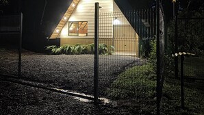 Una casita pequeña de forma triangular con techo inclinado, rodeada por una cerca de alambre y un camino de grava.