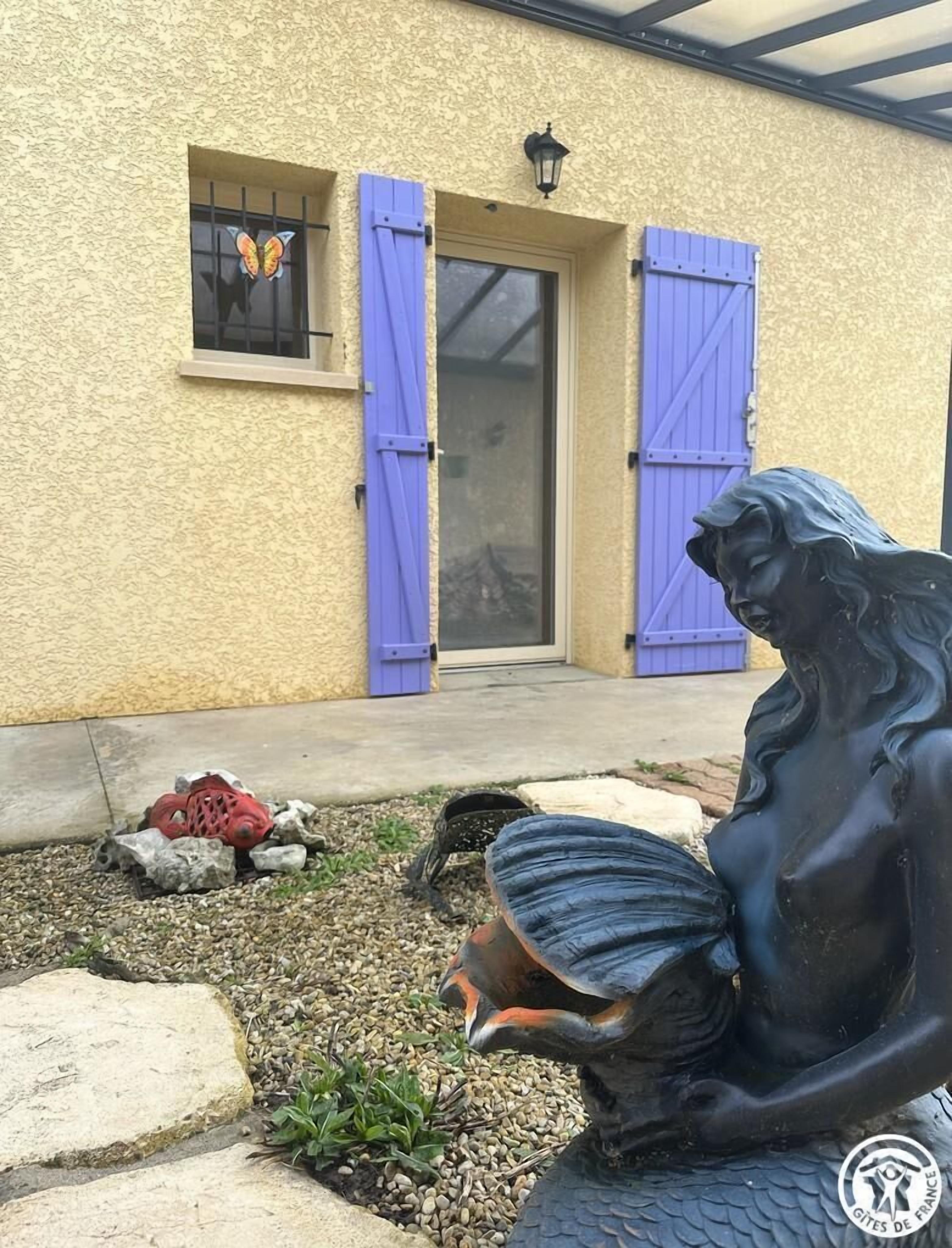 Una estatua de una figura sentada con un velo, ubicada frente a un edificio de enlucido amarillo y contraventanas azules.