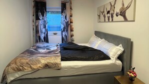 A bedroom with a bed, a cowhide rug, a nightstand, a wall-mounted light fixture, and a painting of animals. - Späth Business Apartment Furnished 2-room basement in Schwanstetten (Schwanstetten)