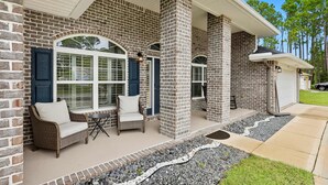 Een huis met een bakstenen gevel, een overdekte veranda met twee rieten stoelen en een tafel, en een pad van stenen dat naar de voordeur leidt.