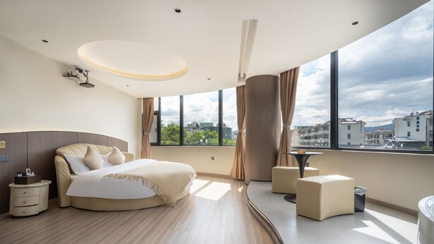 A modern hotel room with a curved bed, a small seating area, and large windows offering a city view. - Ancient City District Ai Shan Hotel (Lijiang)