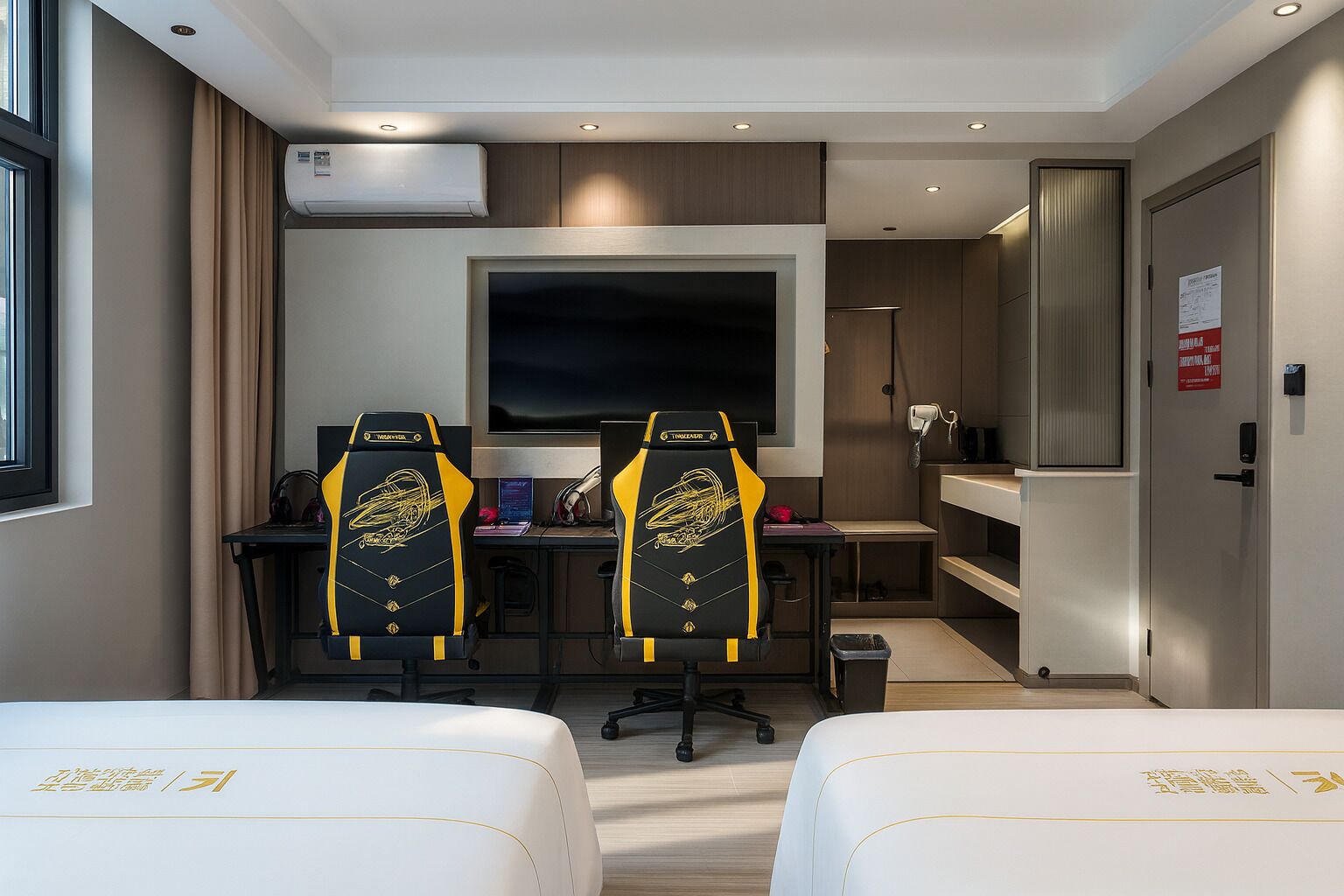 Hotelkamer met twee gamingstoelen, een grote flatscreen-tv, een bureau met een computer, een bed, een kleine kitchenette en een airconditioner.